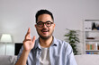 Portrait shot of happy friendly and confident Asian businessman sitting on a couch at home, looking and talking at camera and waving hand saying hello during video call