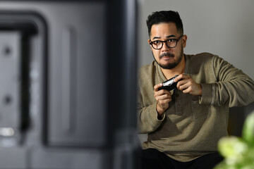 Feeling tense and excited. Young Asian man engaging and playing video game with concentrated expression while sitting on the couch at home.