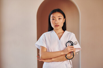 Wall Mural - Doctor with stethoscope. Young serious asian woman standing indoors