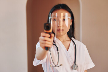 Wall Mural - Holds infrared thermometer. Young serious asian woman standing indoors