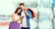 © vgstudio - Holiday sales, shop rebates, consumer credit concept - happy couple with shopping bags, looking at mobile phone. Man and woman over blurred modern interior or mall background.