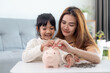 © FAMILY STOCK - Family Savings. Mother Teaching Little Daughter How To Save Money Holding Piggybank