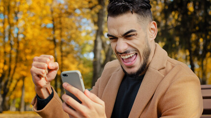 Excited hispanic guy reading good news on smartphone sitting on bench in autumn park young man happily surprised to receive message overjoyed male winner celebrating victory holding telephone outdoors