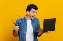 Amazed Happy Excited Confident Unshaven Caucasian Guy, Take Off Glasses, Holding An Open Laptop In His Hand, Looks Surprised And Happily At The Screen While Standing Against Isolated Orange Background