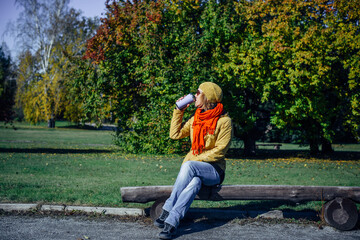 Wall Mural - Pretty european woman in yellow jacket and red scarf sitting on wooden bench in a deserted park and drinking coffee. Enjoying solitude and sunny autumn weather. Blurred plant background, copy space.