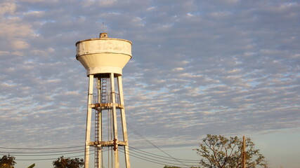 White Concrete Water Tank on Tower: An old public water tank outdoors in the afternoon sunshine with wires and trees below on a gray cloudy sky background with copy space. Selective focus