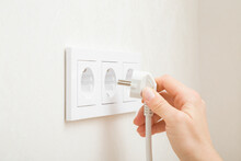 Young Adult Woman Hand Holding And Plugging White Electrical Plug In Wall Outlet Socket At Home. Closeup. Side View.