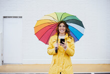 Happy Woman Holding Umbrella And Smart Phone In Front Of Wall