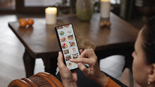 Ordering Food Using A Smartphone At Home. A Woman Selects Sushi Sets In The Internet Menu Of A Japanese Restaurant Using An Application On A Smartphone. Home Evening Furnishings.