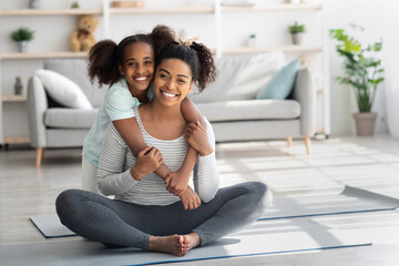 Wall Mural - Sporty black mother and daughter sitting on fitness mat, embracing