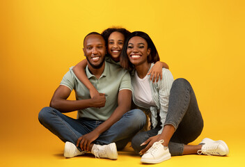 Wall Mural - Family portrait. Happy african american parents and their daughter hugging and smiling, sitting on yellow background