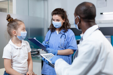 Wall Mural - African american pediatrician doctor analyzing disease symptoms expertise using tablet discussing healthcare treatment with asisstant in hospital office. Medical team with face mask against covid19