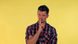 Close up of young man making a hush gesture on yellow background. It must be quite there.