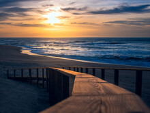 Landscape Of Golden Sunset At The Beach Showing Blurry Close-up Of Illuminated Wooden Handrail Of Boardwalk Leading Down To The Sand And Atlantic Ocean At Quiaios Beach, Portugal