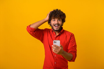 Curly brunette man with beard expressing amazement and holding head with his hands, while using cellphone, with open mouth and shocked big eyes. Indoor studio shot isolated on orange background