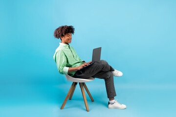 Side view of african american student guy using laptop, surfing internet while sitting in chair on blue background