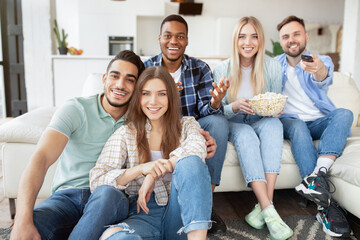 Group of positive young friends enjoying movie at home, eating popcorn, switching channels with remote control