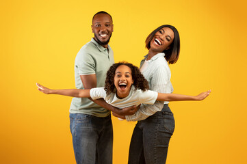 Family bonding concept. Joyful black man and woman holding daughter on hands, having fun over yellow studio wall