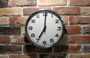 Wall office clock on a brick wall. Seven o'clock. Round clock with minute and hour hands and large numbers in a metal case. Time for getting up in the morning or returning home in the evening.