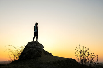 Wall Mural - Silhouette of a woman hiker standing alone on big stone at sunset in mountains. Female tourist on high rock in evening nature. Tourism, traveling and healthy lifestyle concept.
