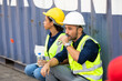 © NVB Stocker - Tired hispanic male and female container worker in hardhat and uniform resting at container yard port of import and export.