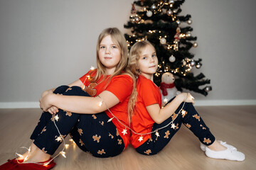 Wall Mural - Teenage sisters in red holiday T-shirts sitting at the Christmas tree and celebrating the New Year