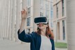 Cheerful redhead female dressed formally stands outdoors in virtual reality goggles