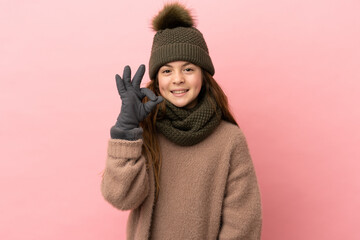 Wall Mural - Little girl with winter hat isolated on pink background showing ok sign with fingers