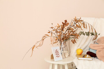 Wall Mural - Armchair with warm clothes, book and vase with autumn branches on table near light wall