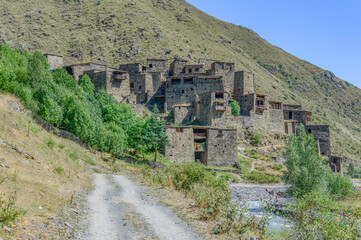 The Fortified Village of Shatili, Khevsureti, Georgia. Abandoned towers and houses, brown and grey old stone walls, green trees and dry grass on mountain slopes, blue sky with clouds, road and river