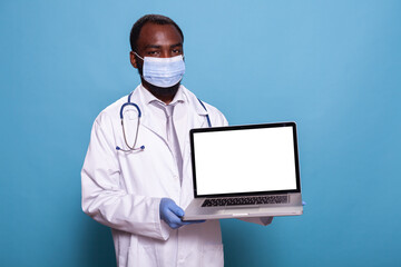 Doctor wearing medical protective gear presenting laptop with white screen mockup for health care concept. Medic in lab coat with stethoscope, face mask and latex gloves holding portable computer.