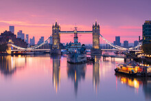 Sunrise View Of HMS Belfast And Tower Bridge Reflected In River Thames, With Canary Wharf In Background, London