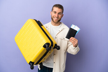 Young handsome caucasian man isolated on purple background in vacation with suitcase and passport