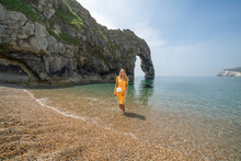 Attractive Woman In Orange Dress Cooling Off On Holiday, Paddling In The Sea By The Durdle Dor, Dorset, England, UK.