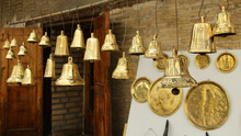 Golden Bells Of Various Sizes Made In The Workshop Of Uzbekistan Hang For Sale Against The Background Of A Brick Wall.