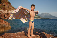 A Little Boy Stands On A Stone On The Beach With A Towel Developing Like Wings In The Wind Against The Backdrop Of The Sea And Rocks.