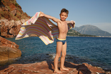 a little boy stands on a stone on the beach with a towel developing like wings in the wind against t
