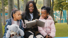 Happy African American Family Resting On Bench In Park Young Mother Reads Book To Daughters Child Holding Teddy Bear Little Girl Points Finger Textbook Mom Smiling Happily Together Spend Time Outdoors