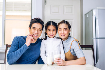 Portrait of enjoy happy love asian family father and mother with little asian girl smiling and having protein breakfast drinking and hold glasses of milk at table in kitchen