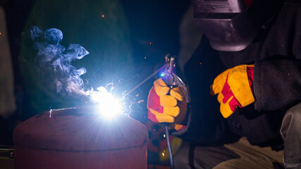 A welder in a workshop with a welding machine, welds metal structures. A worker in a factory uses a welding mask, tools and metalworking equipment.