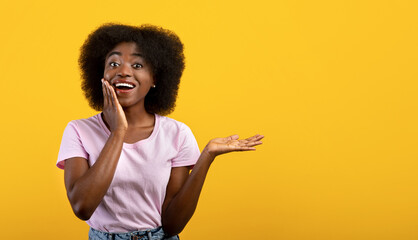 Wow, cool offer. Excited black woman holding hand, showing free space on palm and touching cheek, yellow background