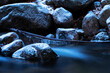 Blue hour, a photo of a mountain stream in the Carpathians in Poland. A huge mass of stones covered with snow and ice. The flowing water turns white and blue. Stone-based branch with ice crystals