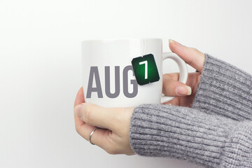 August 7th. Day 7 of month, Calendar date. Closeup of female hands in grey sweater holding cup of tea with month and calendar date on teabag label. Summer month, day of the year concept.