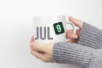 July 9th. Day 9 of month, Calendar date. Closeup of female hands in grey sweater holding cup of tea with month and calendar date on teabag label. Summer month, day of the year concept.