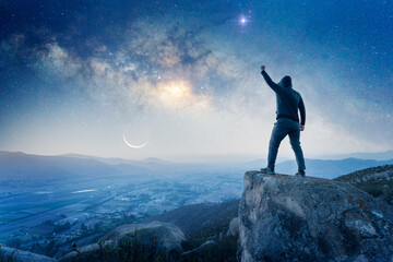 man standing on the top of the mountain, back view, with a hand up celebrating and Milky Way over the valley.