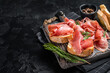 Toast with tomatoes and cured Slices of jamon serrano ham, prosciutto crudo parma on wooden board with rosemary. Black background. Top view. Copy space