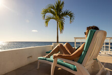 Woman Relaxing Poolside On Vacation Getting A Tan Under The Caribbean Sun With A Dramatic View Of The Ocean From Her Chair On The Tropical Island Of Cozumel In Mexico. 