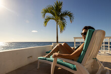 Woman Relaxing Poolside On Vacation Getting A Tan Under The Caribbean Sun With A Dramatic View Of The Ocean From Her Chair On The Tropical Island Of Cozumel In Mexico. 