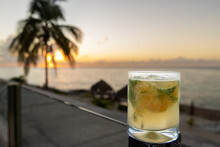 Cocktail Glass With Fresh Made Mojito Featuring Muddled Lime And Mint With A Beautiful View Of Tropical Island Beach Including Sunset And Palm Tree