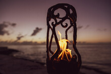 Flames Burn Orange In A Tiki Torch On A Beach In Cozumel, Mexico During Sunset. The Ocean Is In View Behind The Fire.
